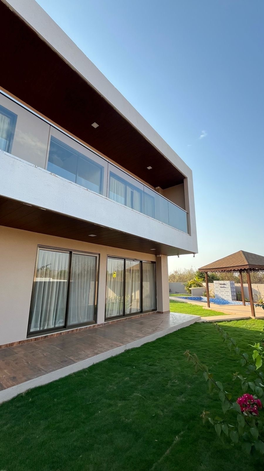 A modern villa exterior is displayed, featuring large glass doors leading to a patio area. The lush green lawn surrounds the property, with a gazebo visible nearby and a private pool in the background, under a clear blue sky.