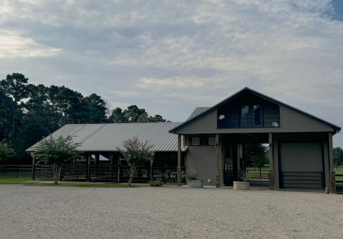 A spacious ranch house is showcased with a metal roof and large open areas. Trees surround the property, and a gravel parking area is visible in front. The entrance includes a wraparound porch, enhancing the welcoming appearance of this peaceful getaway.