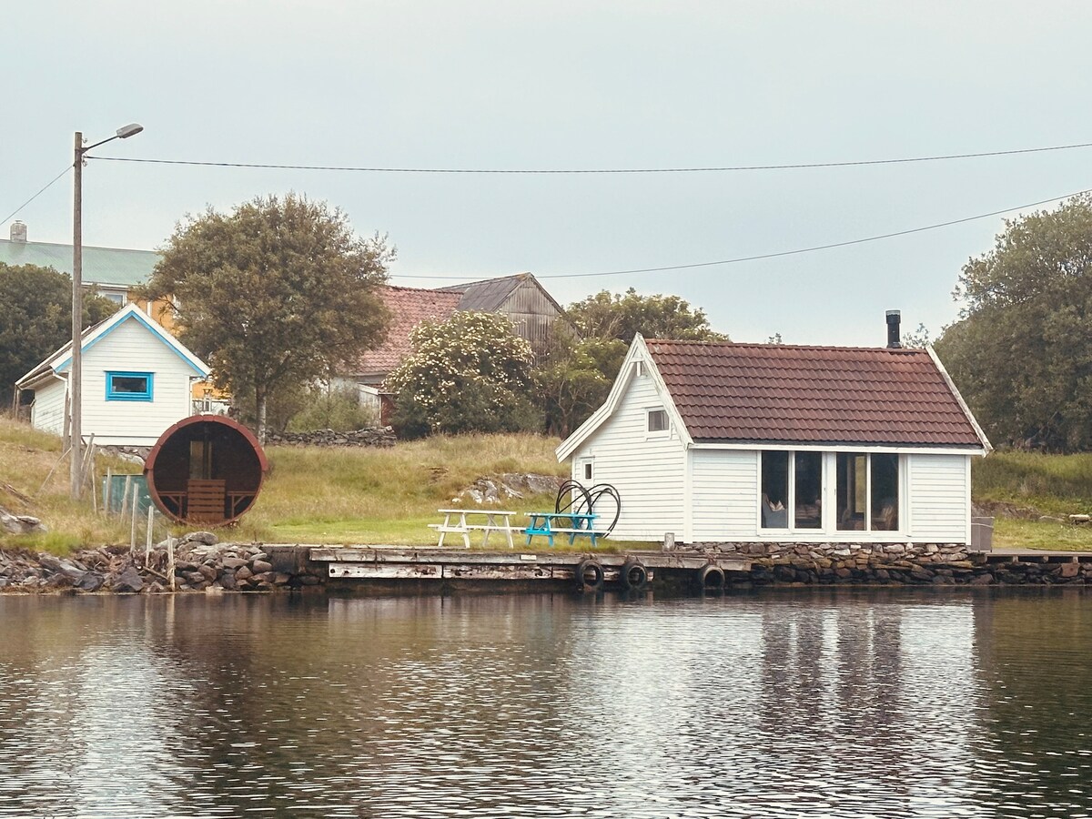 A charming white house sits on the edge of the water, accompanied by a circular wooden sauna nearby. A small outdoor seating area with colorful chairs is visible on the dock, framed by lush greenery and gently sloping land in the background.