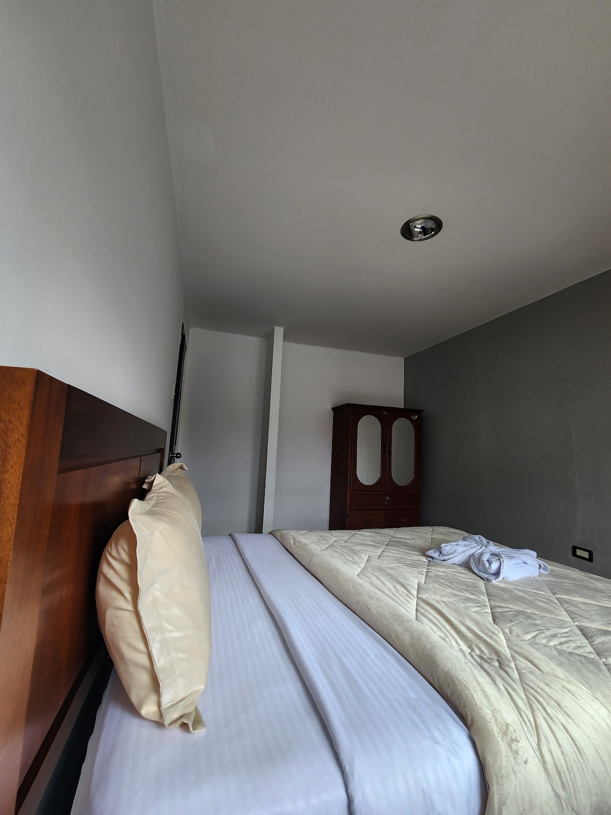 A comfortable sleeping area features a spacious bed with a light-colored blanket. A wooden cabinet stands against the wall, while the room is illuminated by natural light from a nearby source, highlighting the neutral color palette of the walls.