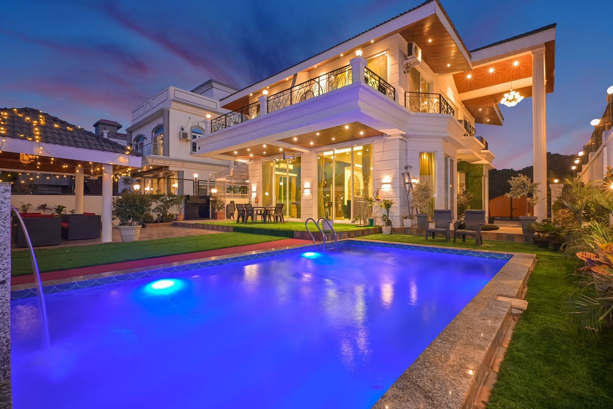 An inviting outdoor pool area features soft blue lighting illuminated at dusk. The spacious villa stands prominently in the background, showcasing multiple levels and large glass doors. Lush greenery surrounds the space, enhancing the serene setting.