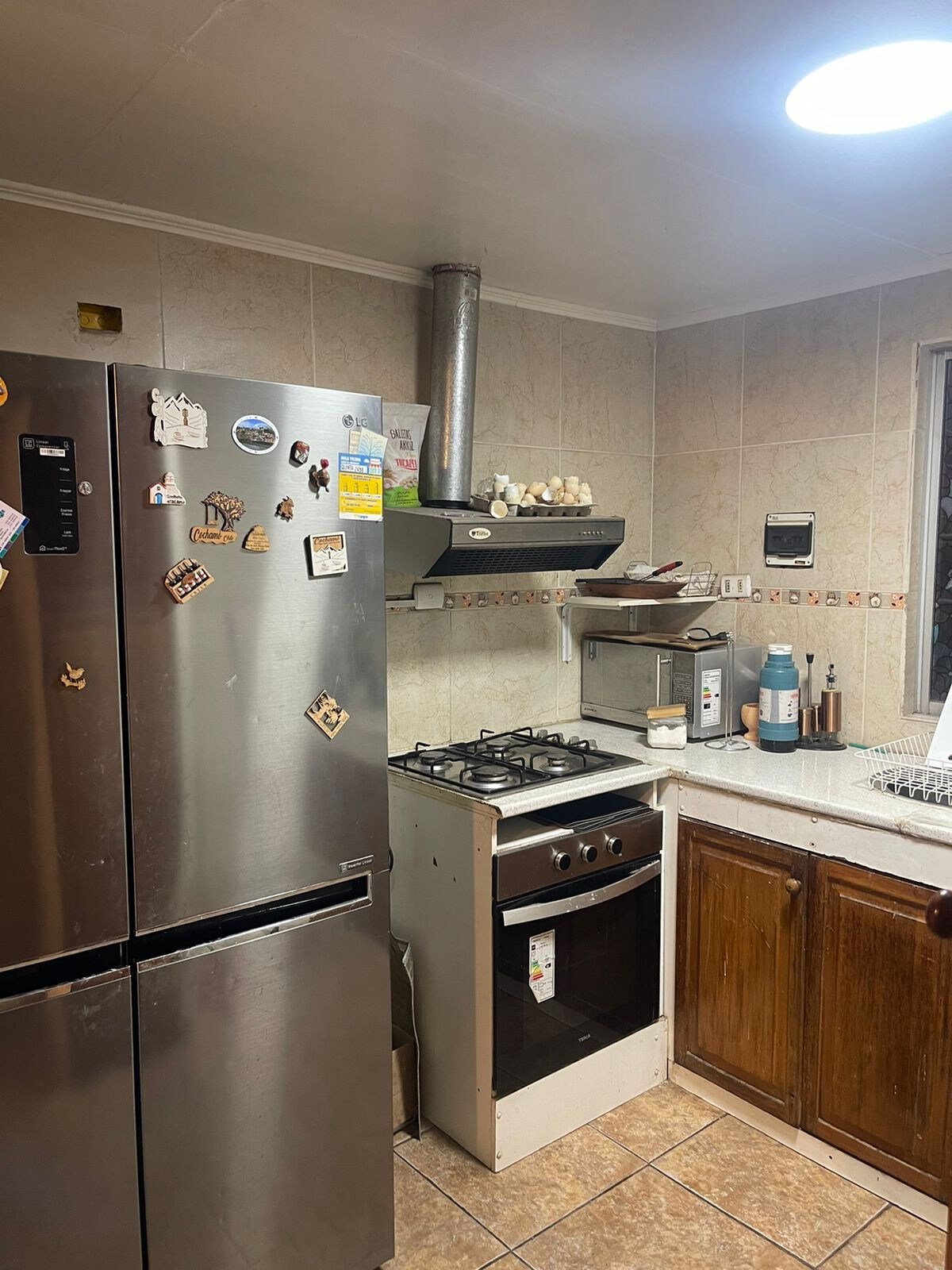 A functional kitchen space is equipped with stainless steel appliances, including a refrigerator and a stove with an oven. Wooden cabinets provide storage, while a countertop offers workspace. Natural light filters in through a window, complementing the neutral tile and wall finish.
