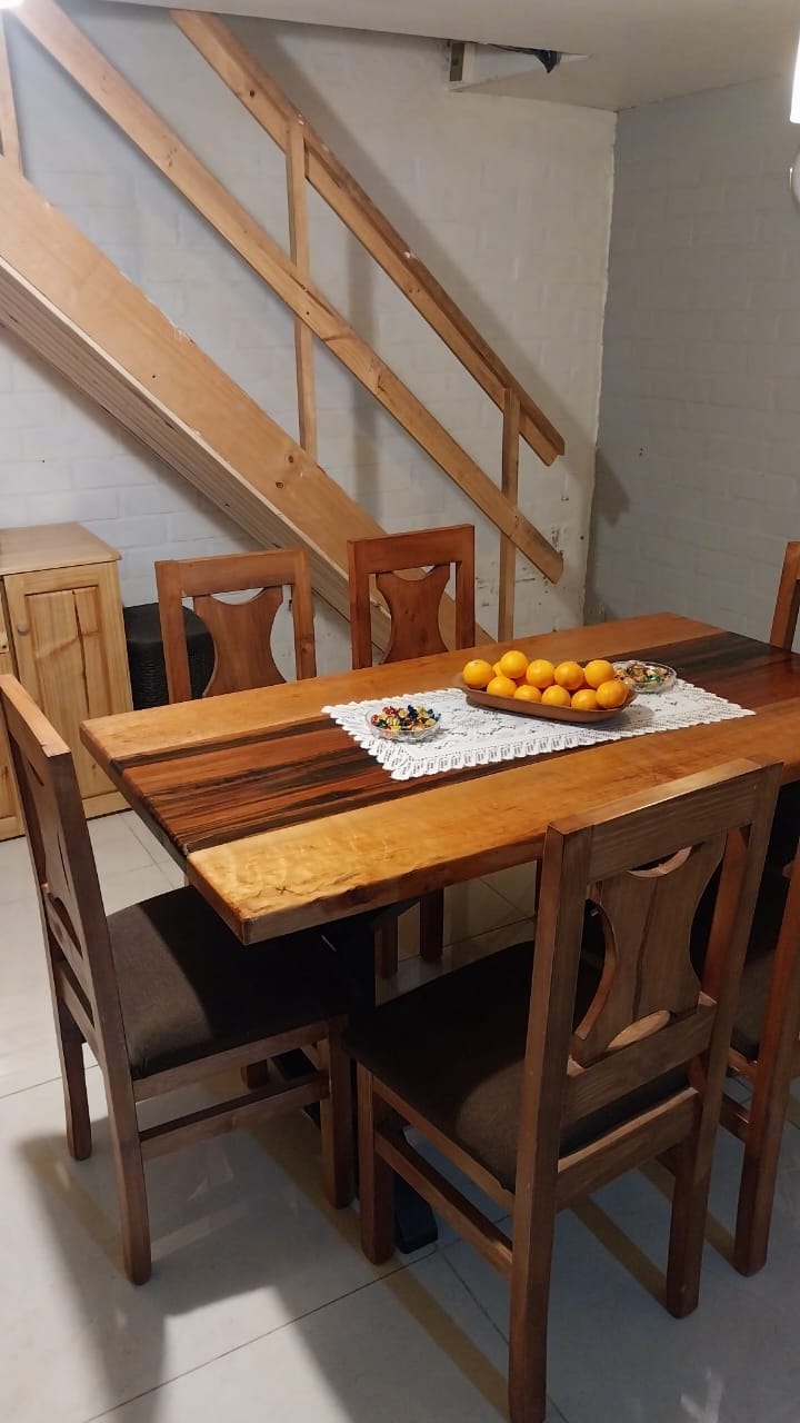 A dining area is presented with a wooden table surrounded by six chairs. A bowl of oranges is center-mounted on a lace doily atop the table. A wooden staircase can be seen in the background, while light reflections play off the tiled floor.