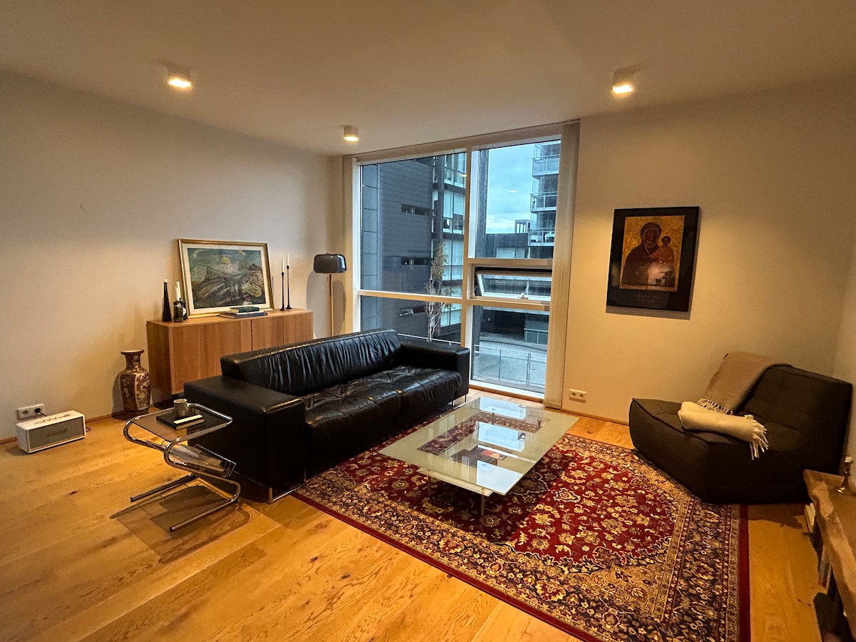 A living area is presented with modern furnishings, including a black leather sofa and a minimalist glass coffee table atop a rich area rug. Large windows allow natural light to enter, with artworks displayed on the walls, creating a refined and inviting environment.