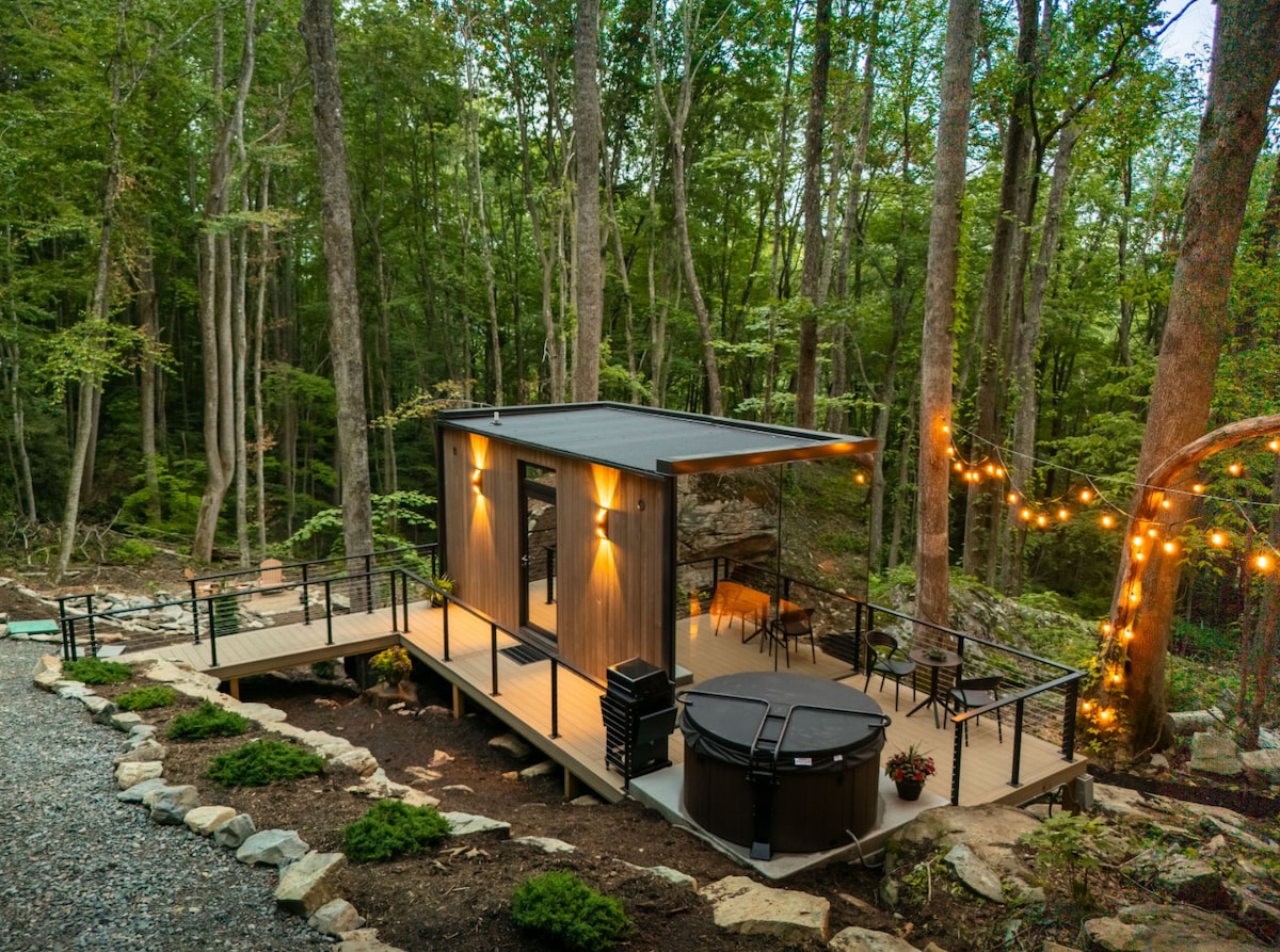 A modern tiny cabin is nestled among trees, featuring a spacious outdoor deck with a hot tub. Soft lighting illuminates the cabin's exterior, while comfortable outdoor seating and dining options are visible on the deck, surrounded by natural landscaping and boulders.