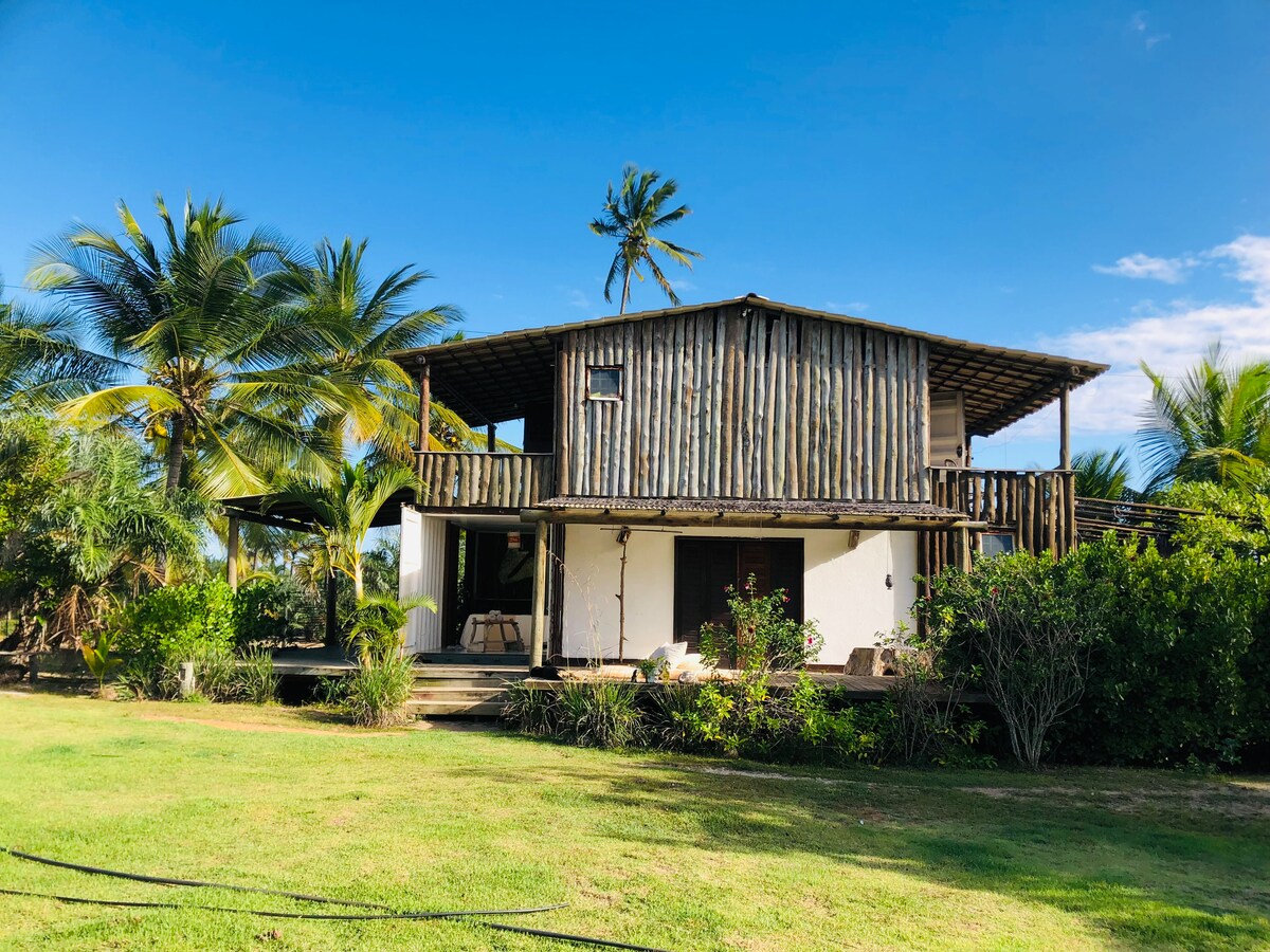 A rustic two-story house constructed with natural materials is surrounded by lush palm trees and greenery. A spacious outdoor deck is visible, offering a relaxing area with comfortable seating. The bright blue sky adds a serene backdrop to the peaceful landscape.