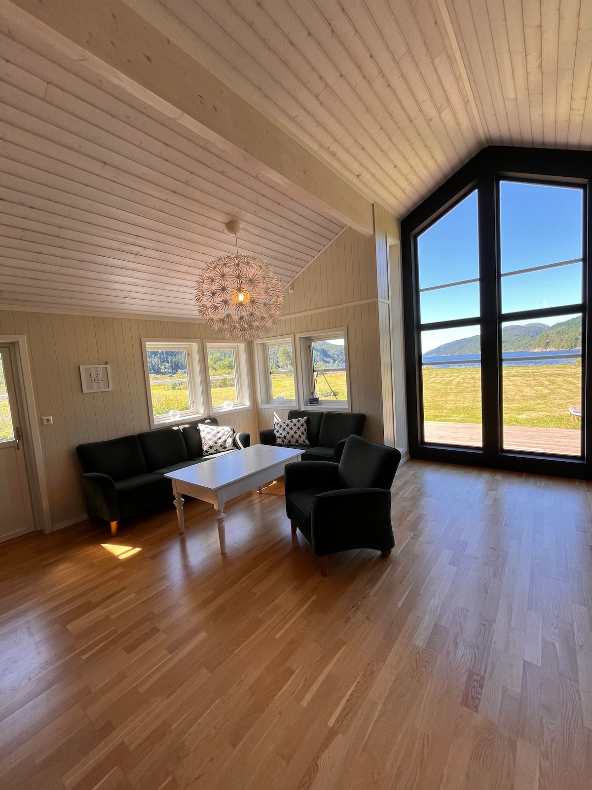 A bright living area is furnished with two comfortable black armchairs and a white coffee table. Tall windows offer a view of the serene fjord, with natural light illuminating the wooden floors and highlighting the unique chandelier above.