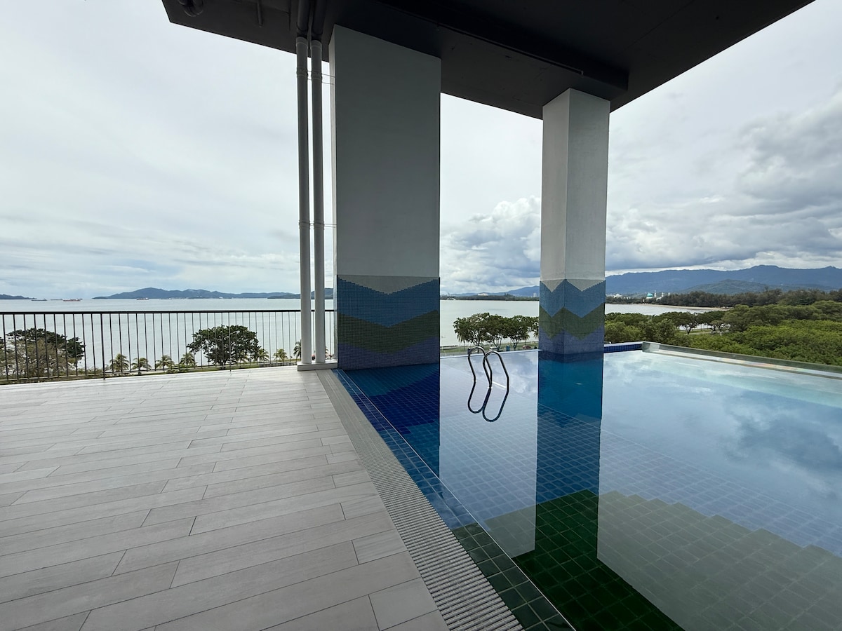 An infinity pool extends towards the ocean, with steps leading into the water. The pool's tiled surface features shades of blue and green, reflecting the surrounding landscape. The backdrop includes distant hills under a cloudy sky, enhancing the serene atmosphere.