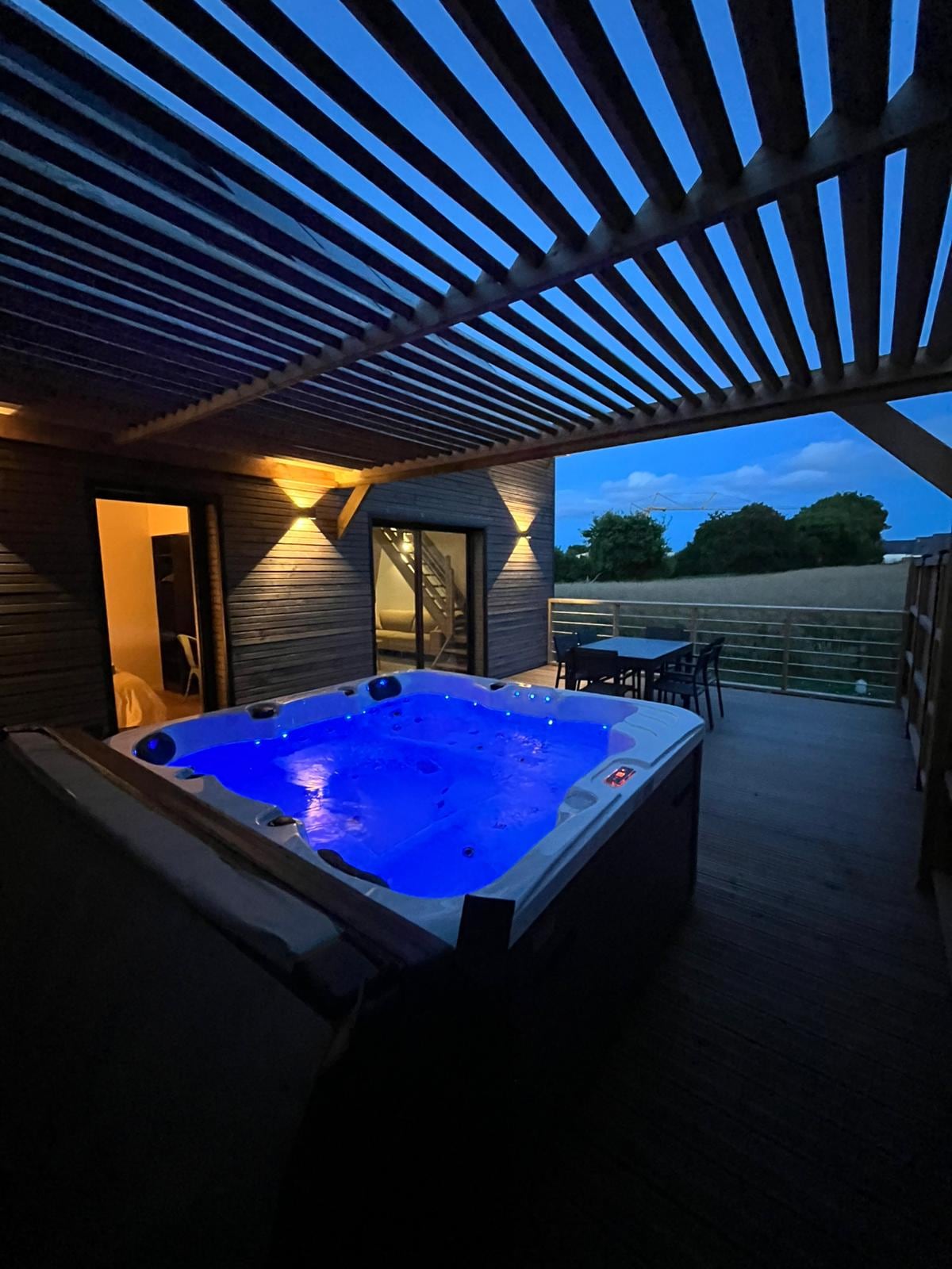 A private hot tub is illuminated with soft blue lighting on a covered terrace. Wooden slats above provide shade, while a small dining area with a table and chairs is visible nearby. Gentle outdoor lighting enhances the serene atmosphere during twilight.