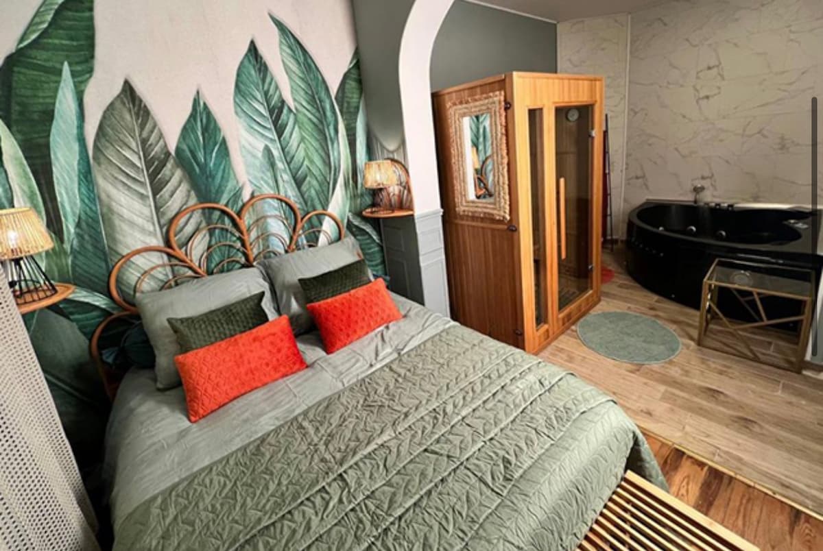 The room features a spacious bed with green bedding and accent pillows in shades of orange. A large mural of tropical leaves adorns the wall behind the bed. To the right, a sauna is visible, with a black whirlpool bathtub beyond it, complementing the warm wood and stone textures.