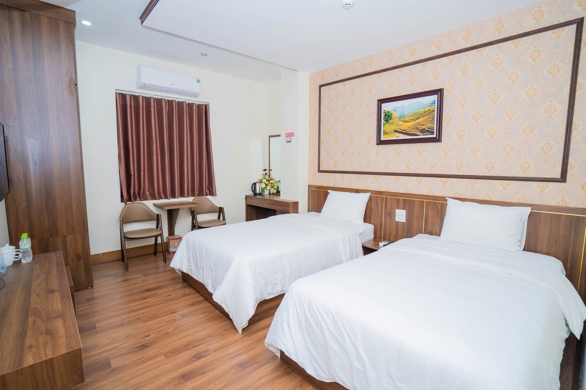 A room with two single beds is furnished with light-colored linens. A wooden desk and two chairs are positioned near a window with brown curtains. A flat-screen TV is mounted on the wall opposite the beds. A painting hangs above the beds, adding a subtle decorative touch.