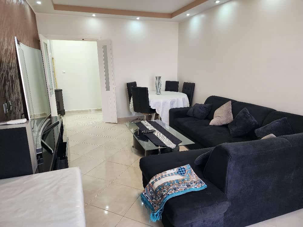 A functional living space features a dark sectional sofa and a glass coffee table, complemented by decorative cushions. A circular dining table with four chairs is situated nearby. The room is illuminated by soft ceiling lights, creating a welcoming atmosphere. A light-colored accent wall enhances the overall brightness.