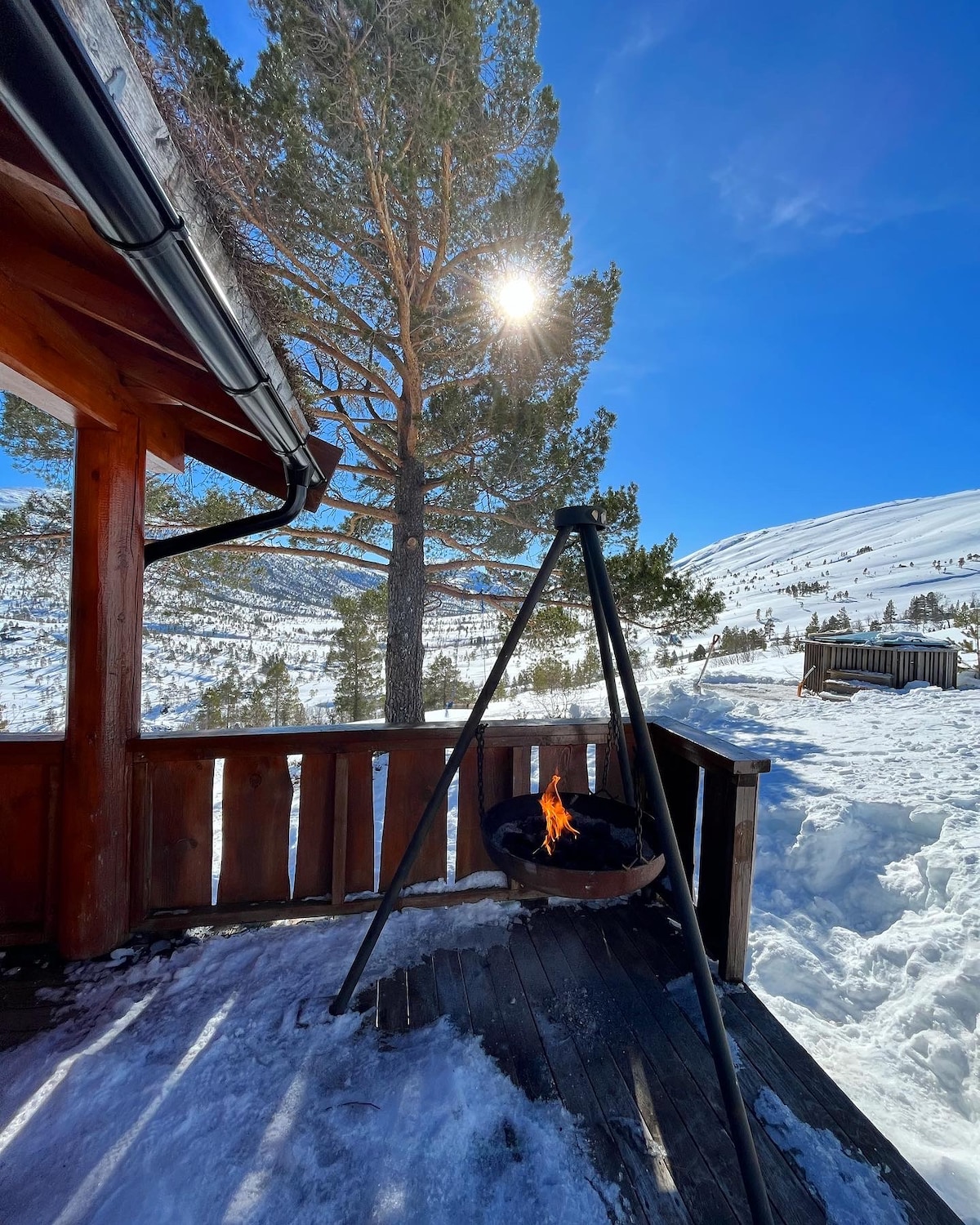 A wooden veranda extends outward, featuring a tripod fire pit with flames visible. A large tree stands nearby, with sunlight casting clear shadows on the snowy ground. The bright blue sky creates a serene backdrop, showcasing the natural beauty of the surroundings.
