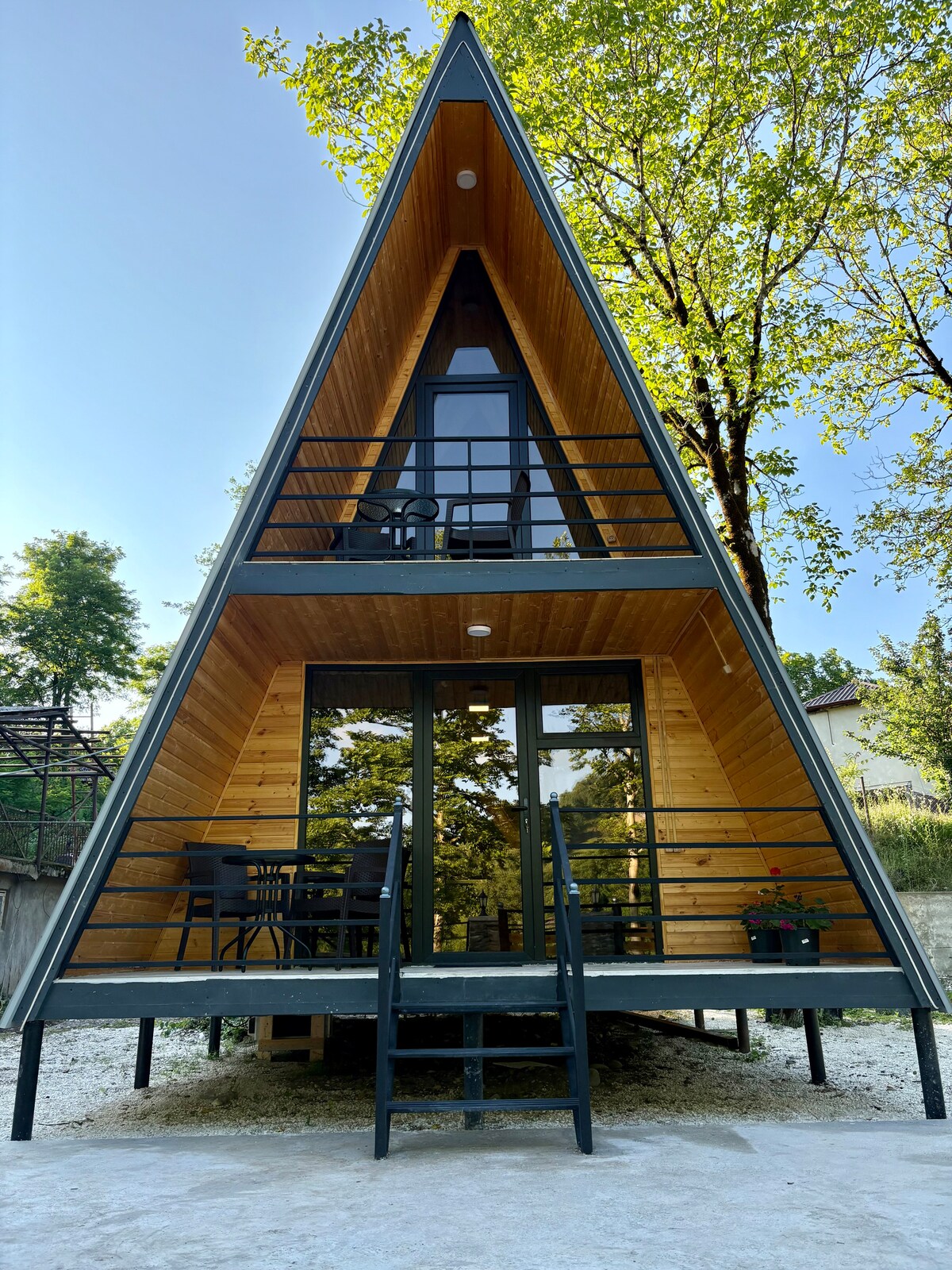 An A-frame cottage showcases a modern design with wooden accents. The entrance features large glass doors and a spacious deck with outdoor seating. Lush greenery surrounds the structure, enhancing the tranquil setting. A staircase leads up to the entrance, inviting guests to enter.