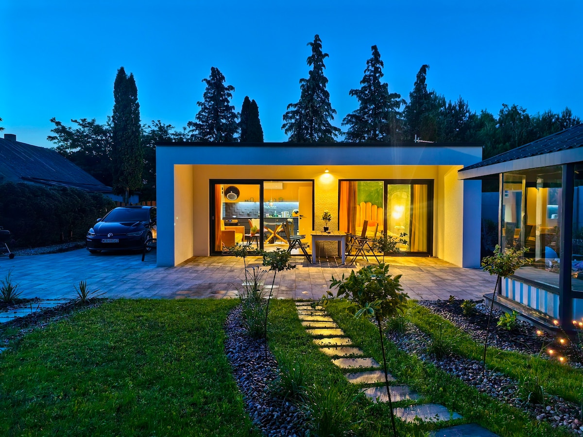 The exterior of a modern house is illuminated by soft outdoor lighting, providing a welcoming glow. Large glass sliding doors open to a terrace with seating, framed by tall trees. A stone pathway leads through a well-maintained garden, enhancing the serene atmosphere of the property.