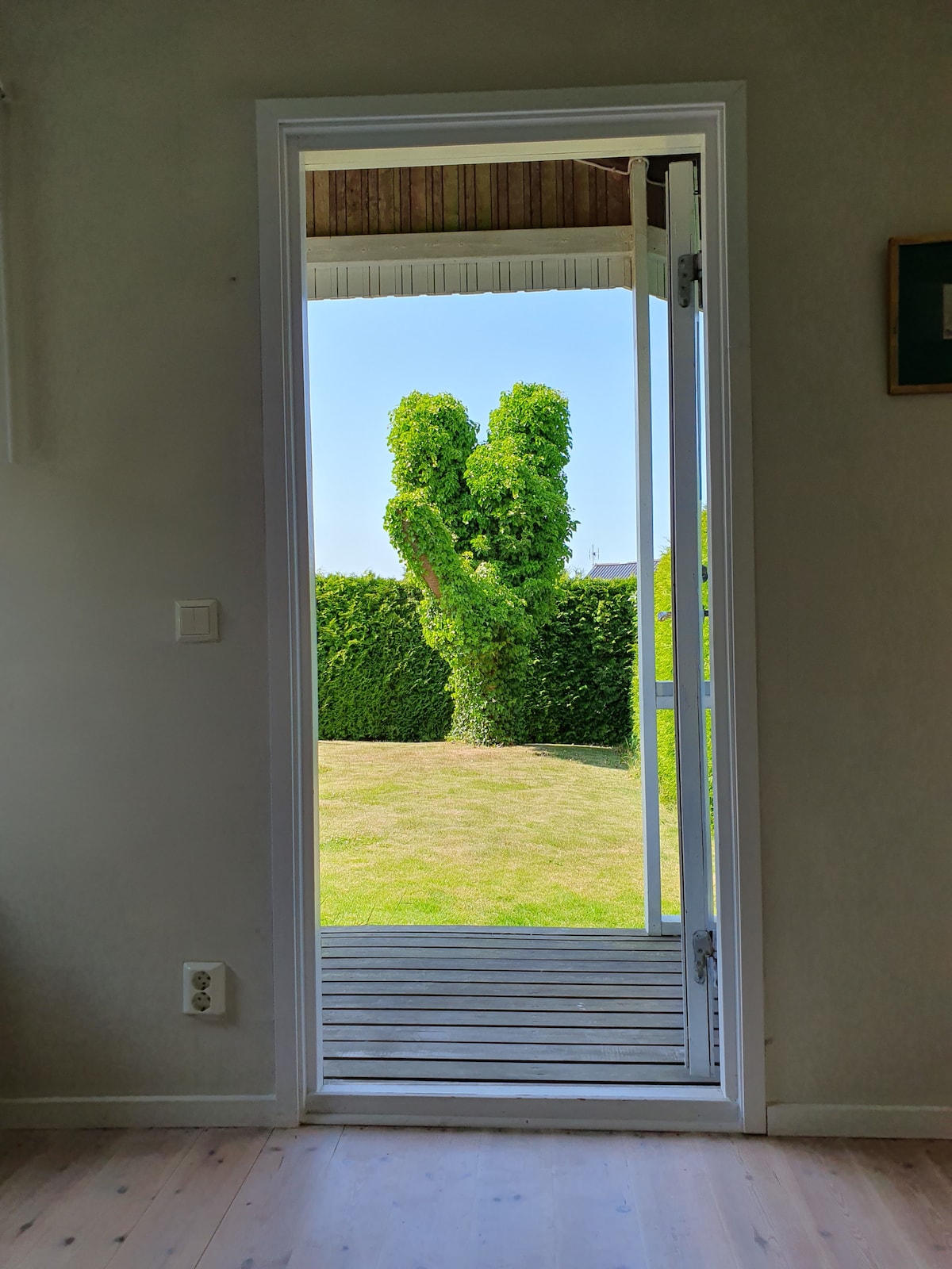 An open door reveals a view of a green garden with neatly trimmed hedges. A small wooden deck is visible in front, providing a connection to the outdoor space. Natural light brightens the interior, enhancing the welcoming entryway.