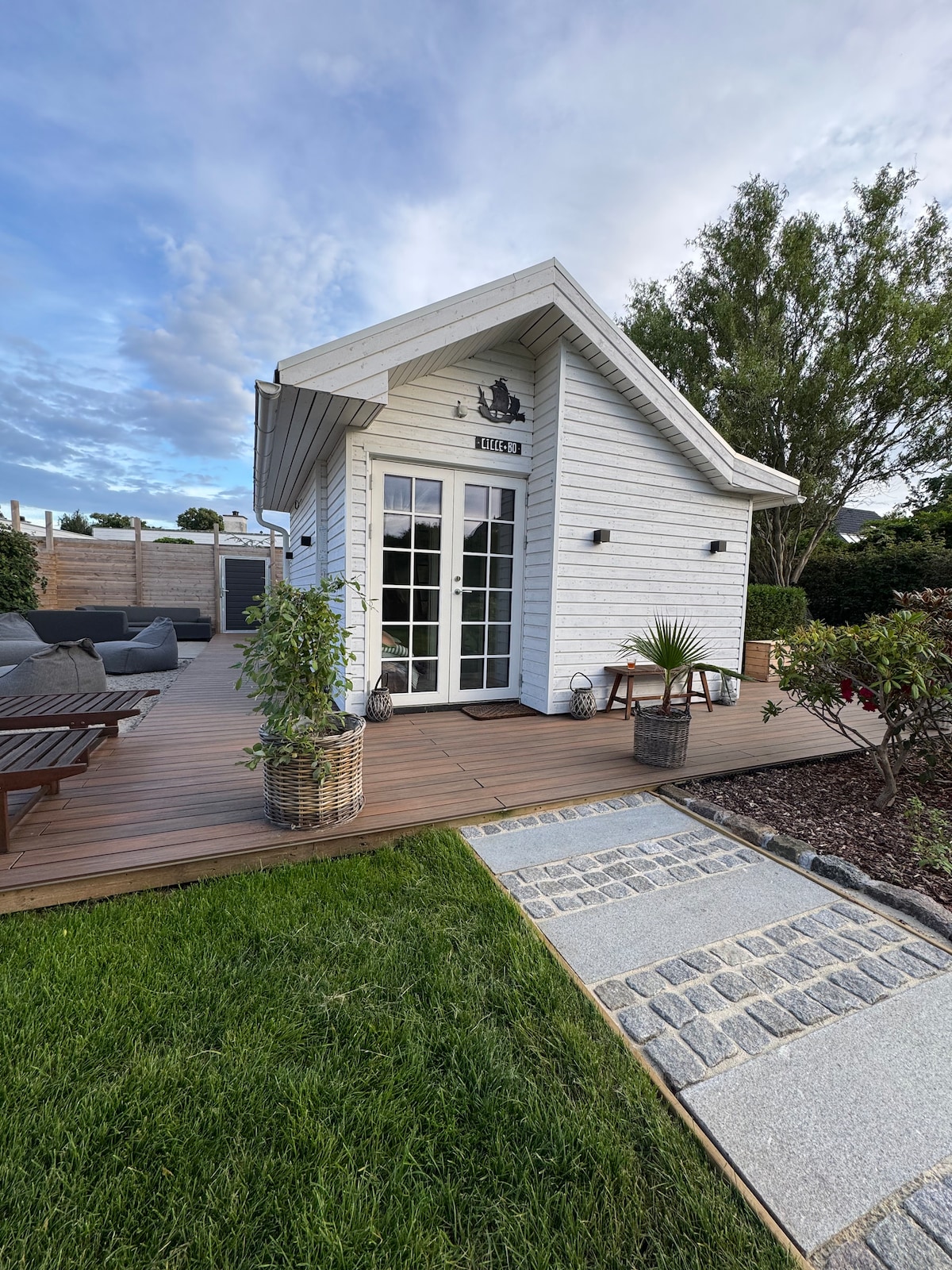 A charming exterior of a small white house features a wooden deck with outdoor seating. Potted plants are placed beside the entrance, leading to a pathway made of light-colored stones. Lush green grass surrounds the area, complemented by a clear sky.