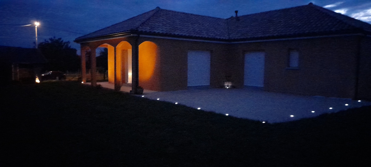 The house features a warm exterior glow, accentuated by lights that outline the entrance. A covered porch offers shaded space, while the surrounding lush grass extends, presenting a sense of openness. The structure’s tiled roof and simple architectural lines contribute to a serene ambiance.