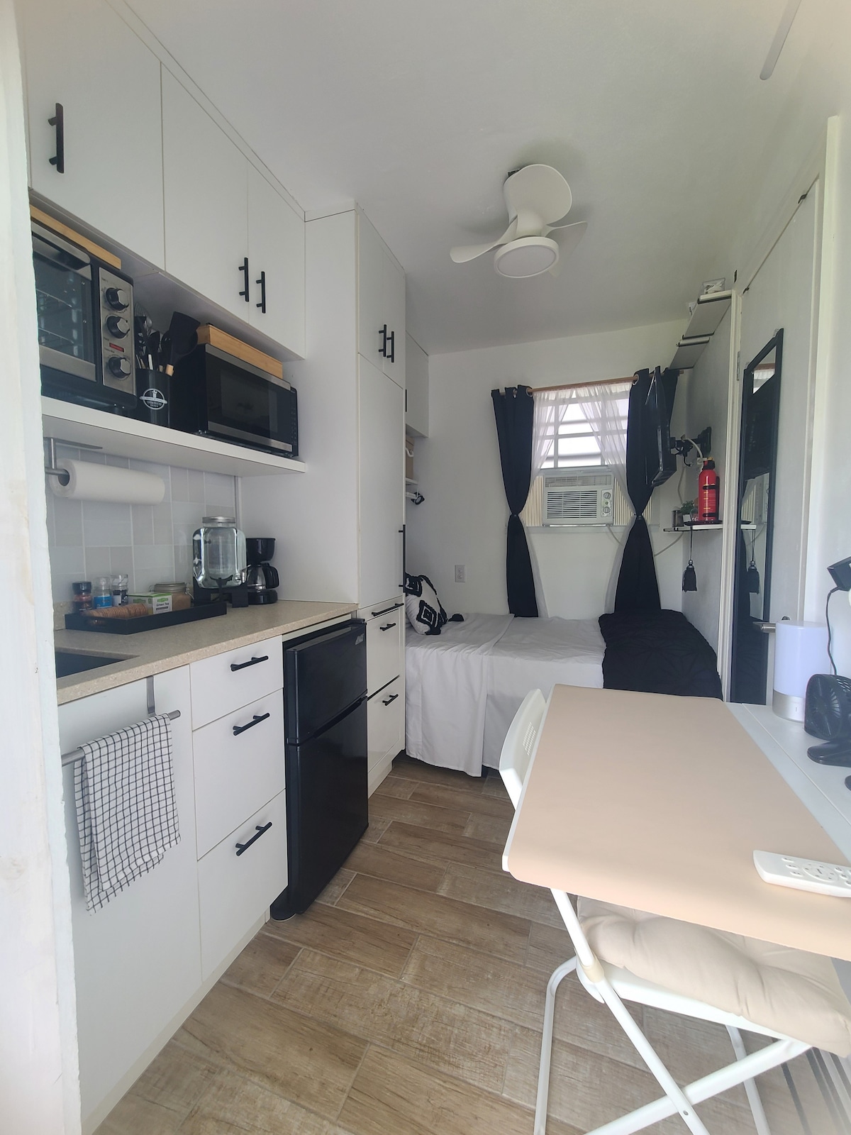 A compact microstudio is shown, featuring modern appliances and cabinetry in a light-toned kitchen area. A versatile workstation is visible next to a cozy sleeping space. Dark curtains frame the window, while a ceiling fan provides air circulation, contributing to the room's functionality.