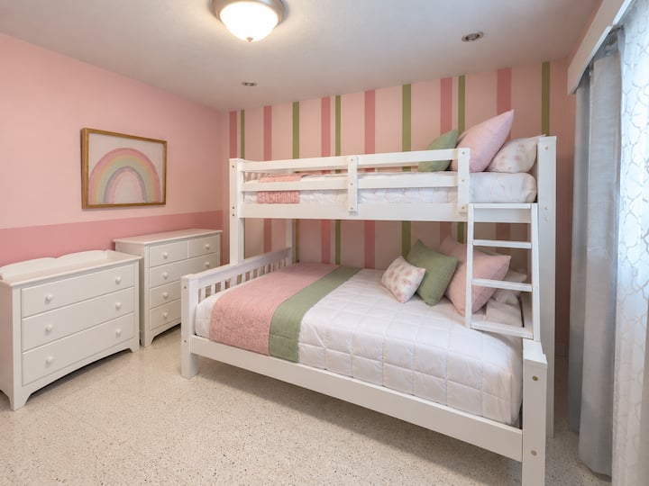 This bedroom features a white twin-over-full bunk bed, providing comfortable sleeping space for up to three guests. Ideal for families traveling with children, the room also includes a playpen for a baby.