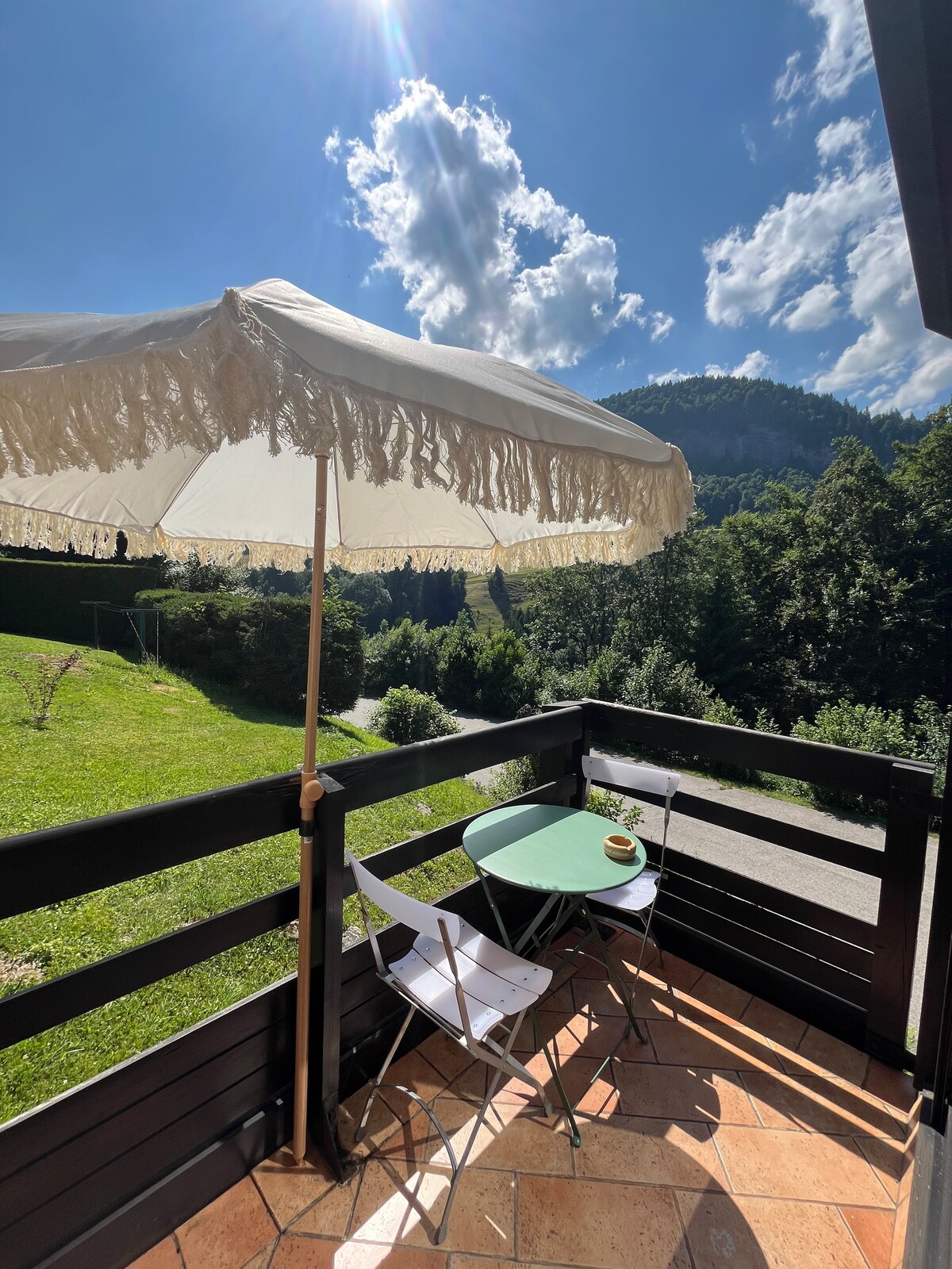 A balcony features a round green table accompanied by two white chairs, sheltered under an elegant parasol. The scenic view includes lush green trees and distant mountains, with a bright sky highlighted by fluffy clouds.