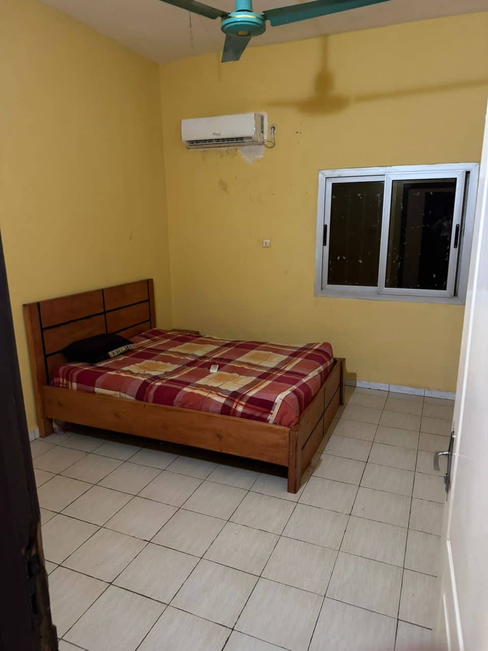 A bedroom features a wooden bed with a plaid blanket and a simple headboard. A window provides natural light, while an air conditioning unit is mounted on the wall. Neutral-colored tiled flooring complements the yellow walls.