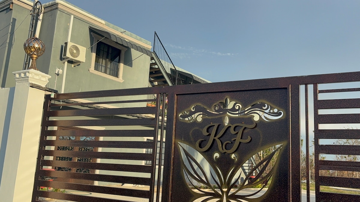 A stylish entry gate features intricate metalwork with the initials 'KF' prominently displayed. The gate is framed by a well-maintained concrete wall, and the upper balcony of the building is visible above, complemented by a spiral staircase leading to it.