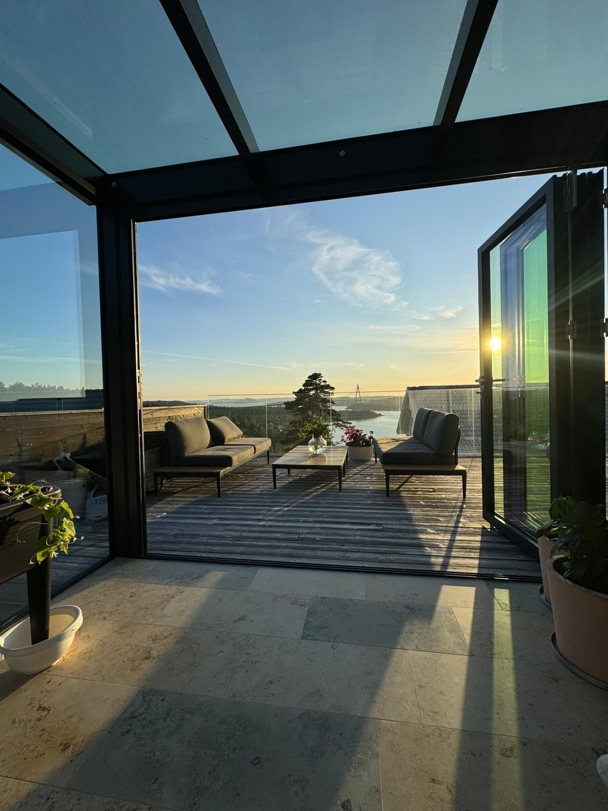 An enclosed terrace is captured, featuring sleek furniture arranged for relaxation. The scene showcases a scenic view of the sunset over the horizon, with glass panels allowing natural light to fill the space. Potted plants are present, adding a touch of greenery.