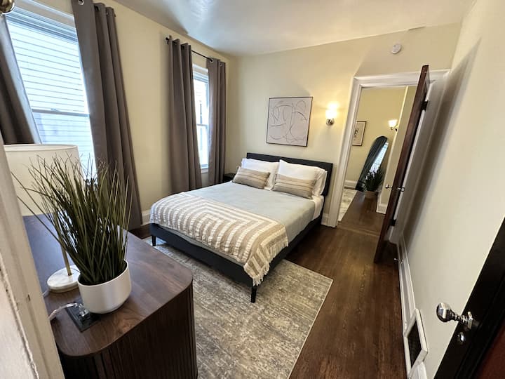 Acogedor dormitorio con una cama tamaño queen, cortinas opacas, cómoda y una mesita de noche con estación de carga integrada. Un espacio cómodo y tranquilo para relajarse después de explorar Búfalo.