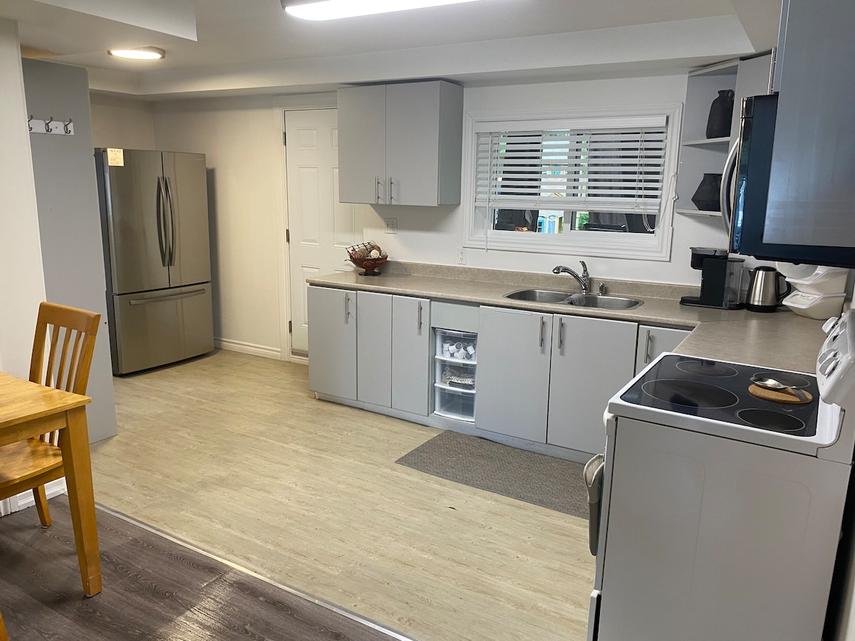 A modern kitchen is arranged with grey cabinetry and a stainless steel refrigerator. A small dining table with wooden chairs is positioned nearby. Natural light enters through the window with white shutters, illuminating the countertop and the various kitchen appliances.