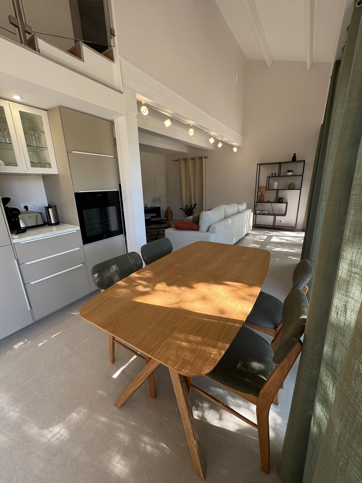 A well-lit dining area is shown, featuring a wooden table surrounded by four chairs. The modern kitchen cabinets are visible in the background alongside appliances. Natural light filters through large windows, casting shadows across the floor, while a cozy living area can be glimpsed in the distance.