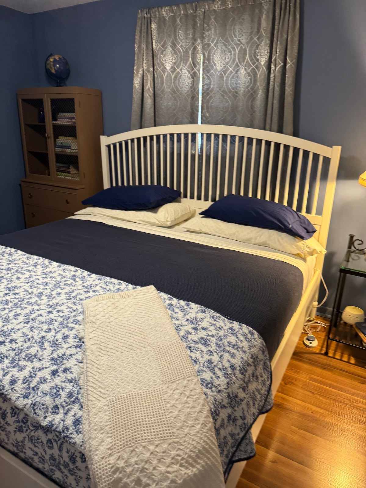 A spacious bedroom features a comfortable king-sized bed with blue and white bedding. A decorative throw lies across the foot of the bed. Natural light enters through a window adorned with sheer curtains, while a wooden cabinet and bedside lamp complete the inviting space.