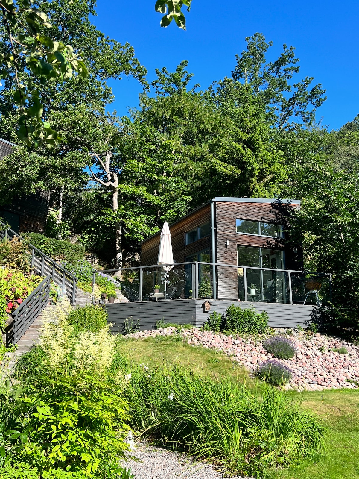 A modern guesthouse is surrounded by lush greenery and trees. The building features large windows and a patio area with an umbrella. A landscaped garden with various plants and stones is visible in the foreground, creating a harmonious connection with the natural setting.