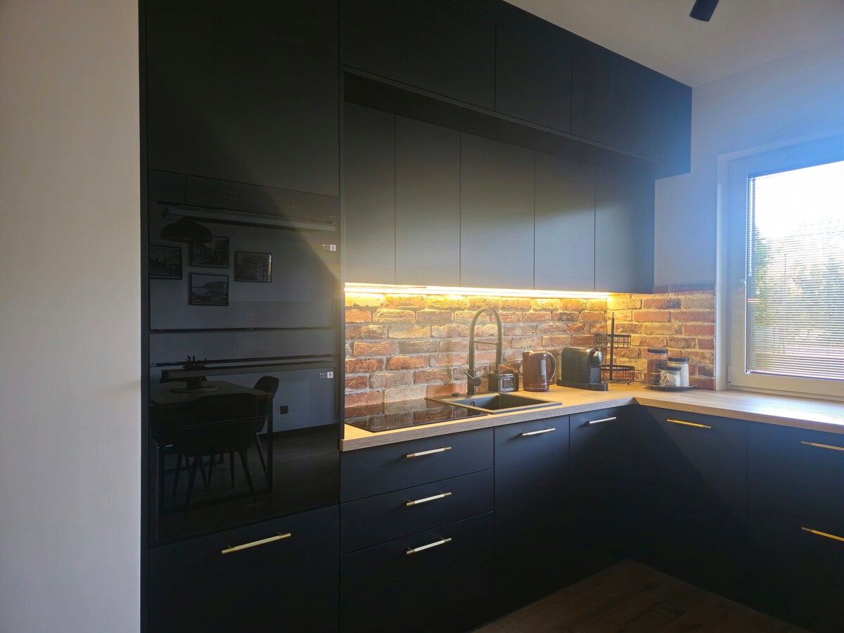 A modern kitchen features sleek black cabinetry and a warm brick backsplash. Under-cabinet lighting illuminates the countertop area, enhancing the workspace. A window allows natural light to enter, providing a view of the outdoors while modern appliances are neatly integrated into the design.
