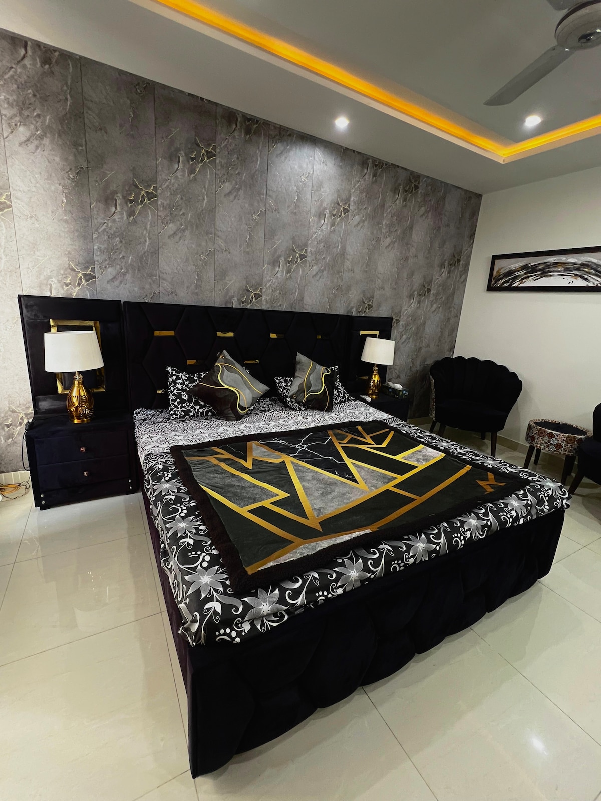 A modern bedroom features a spacious bed with a geometric patterned coverlet in black and gold. Flanking the bed are two elegant bedside lamps. The wall is adorned with textured gray panels, and a stylish armchair adds to the contemporary decor.