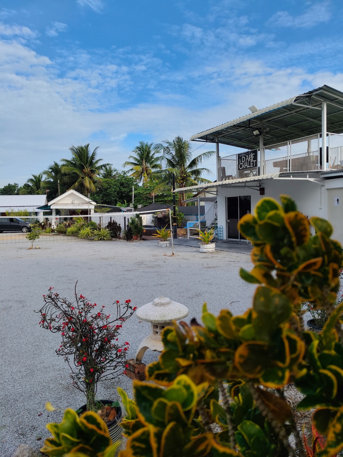 A spacious outdoor area is surrounded by greenery and decorative plants. The building features a modern design with a covered balcony, providing a relaxed environment. Sunshine filters through the palm trees, creating a bright and inviting atmosphere for visitors.