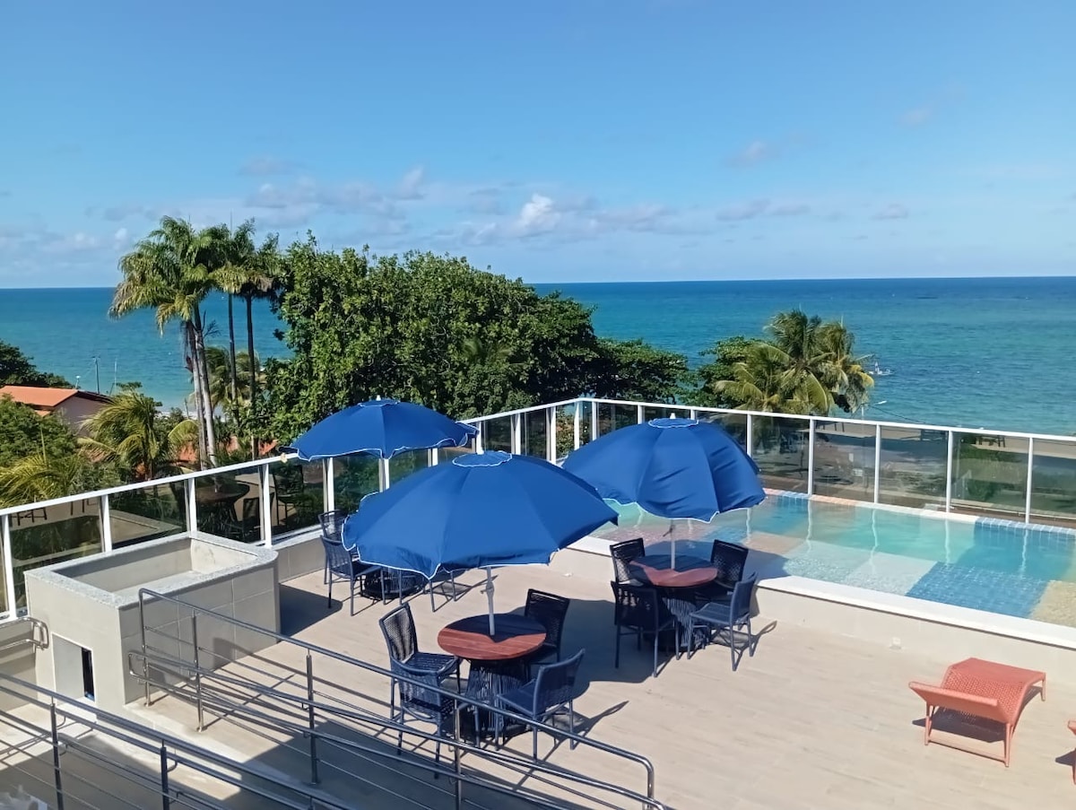 An outdoor deck area is depicted with several blue umbrellas providing shade over patio tables and chairs. The pool is visible in the background, alongside a view of palm trees and the ocean beyond. The scene is filled with natural light and clear skies.