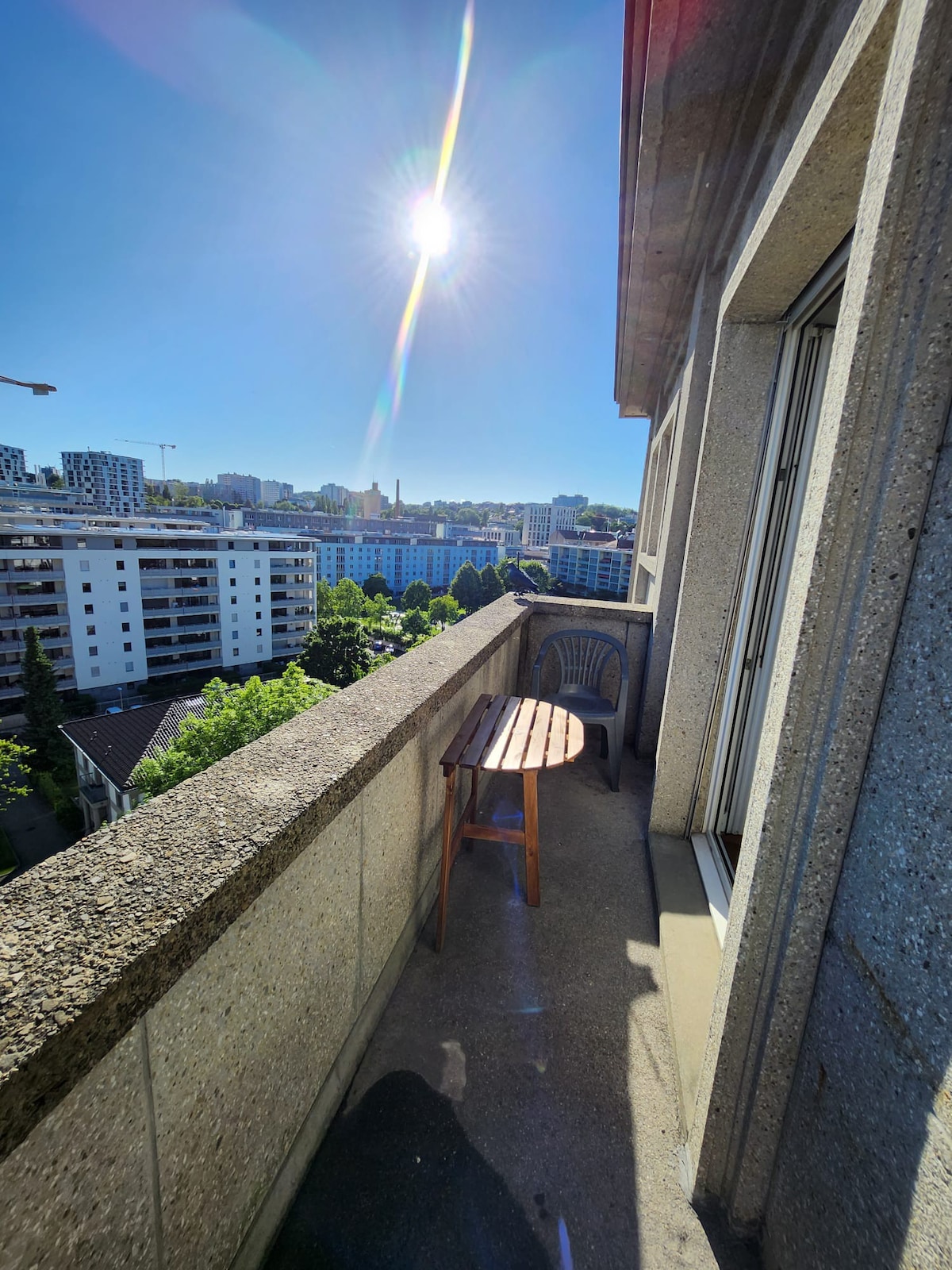 A private balcony extends from the room, featuring a small wooden table and a single chair. The sun shines brightly in a clear blue sky, while city views are visible, showcasing buildings and greenery in the surrounding area.