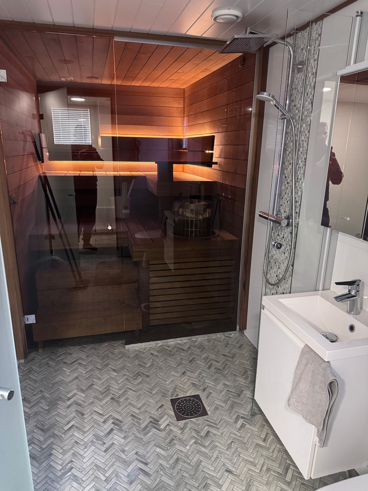 A spacious bathroom features a wooden sauna enclosed by glass, with ambient lighting highlighting the interior. A modern shower is present, along with a sleek sink and storage unit. The floor is designed with patterned tiles, contributing to the bathroom's contemporary look.