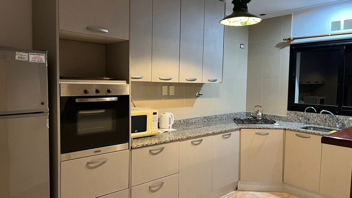 A functional kitchen area features light-colored cabinets and a granite countertop. Modern appliances include a refrigerator, microwave, and oven. A sink is positioned near a window that allows natural light to brighten the space, while dark accents provide contrast.