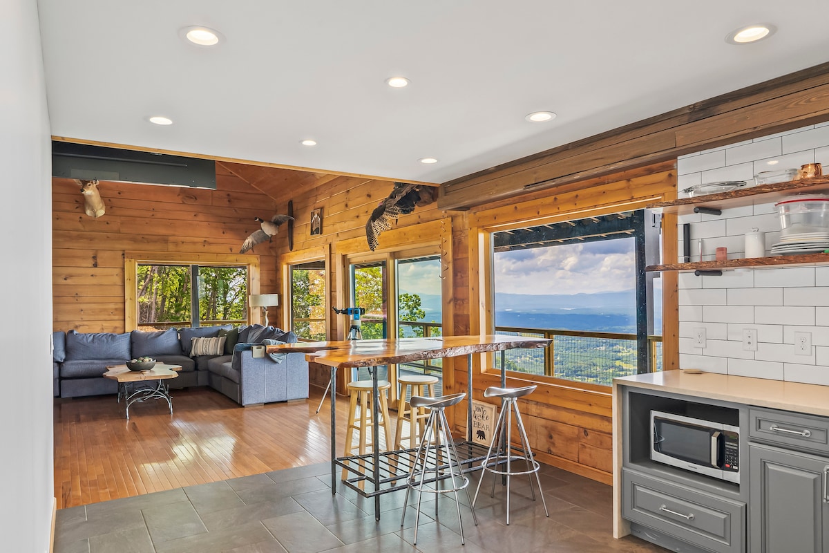 A spacious living area features large windows that frame a scenic mountain view. Natural wood accents complement a sectional sofa and a dining table with metal stools. Light is provided by recessed lighting and the warmth of natural wood beams throughout the room.