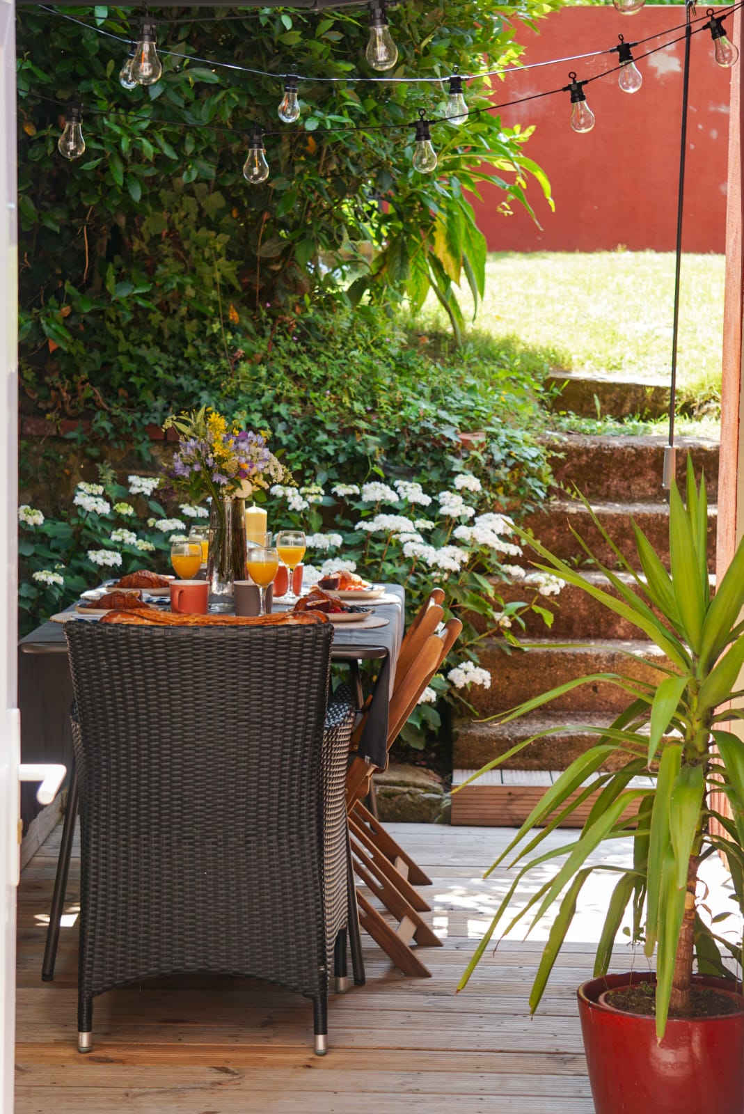 An outdoor dining area features a table set for a meal, surrounded by greenery and white flowers. The setting includes a variety of drinks, along with decorative tableware. Soft lighting from hanging bulbs enhances the inviting atmosphere of the space.