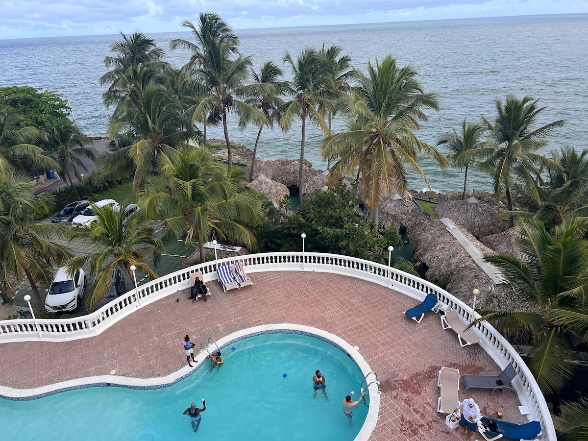 A spacious pool area is surrounded by palm trees, with lounge chairs positioned around its edge. The calm sea is visible in the background, along with thatched-roof huts. Several guests are seen enjoying the pool, enhancing the relaxing atmosphere.