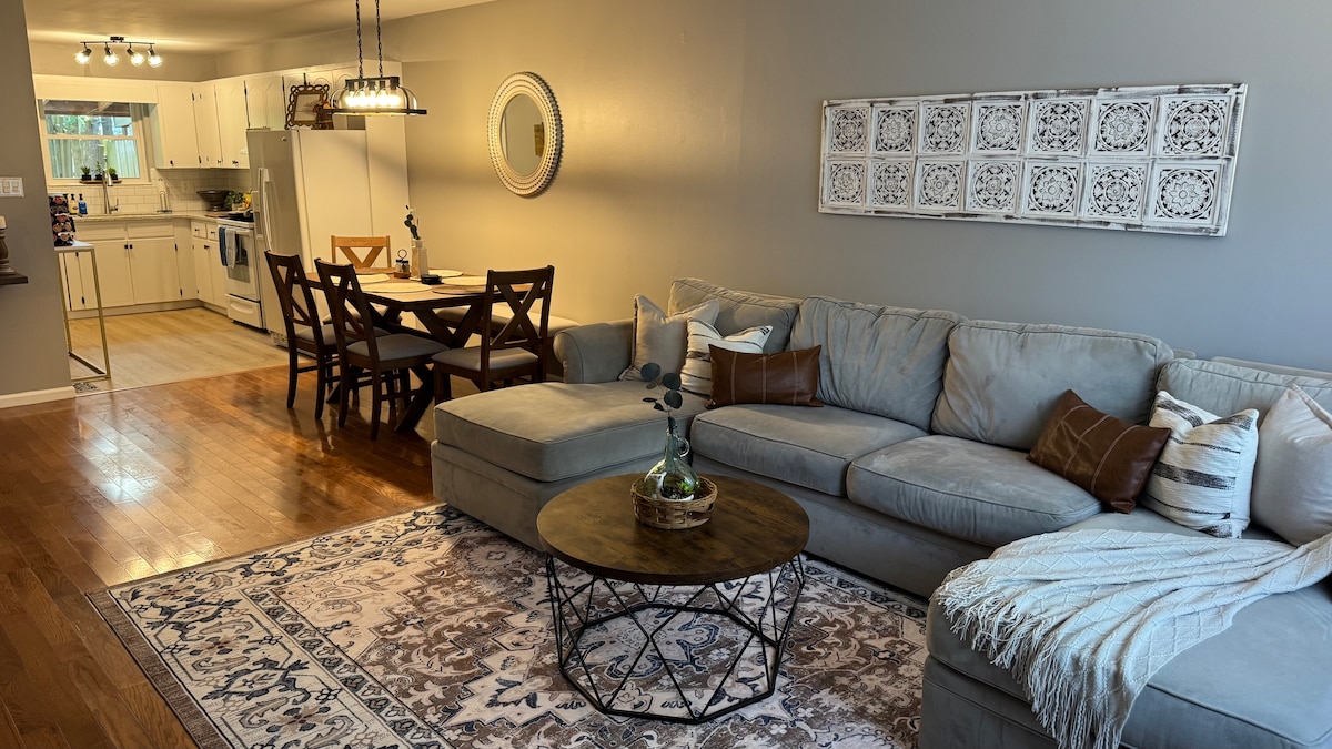 A spacious living area is showcased, featuring a large sectional sofa adorned with decorative pillows. A round coffee table sits at the center, surrounded by a contemporary area rug. In the background, a dining table is visible, accompanied by dining chairs, and a well-equipped kitchen area is glimpsed.