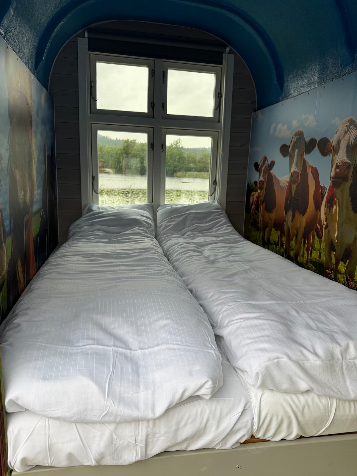 A sleeping area features two cozy beds arranged side by side, dressed in white linens. Large windows overlook a scenic view of water and greenery, allowing natural light to brighten the space.