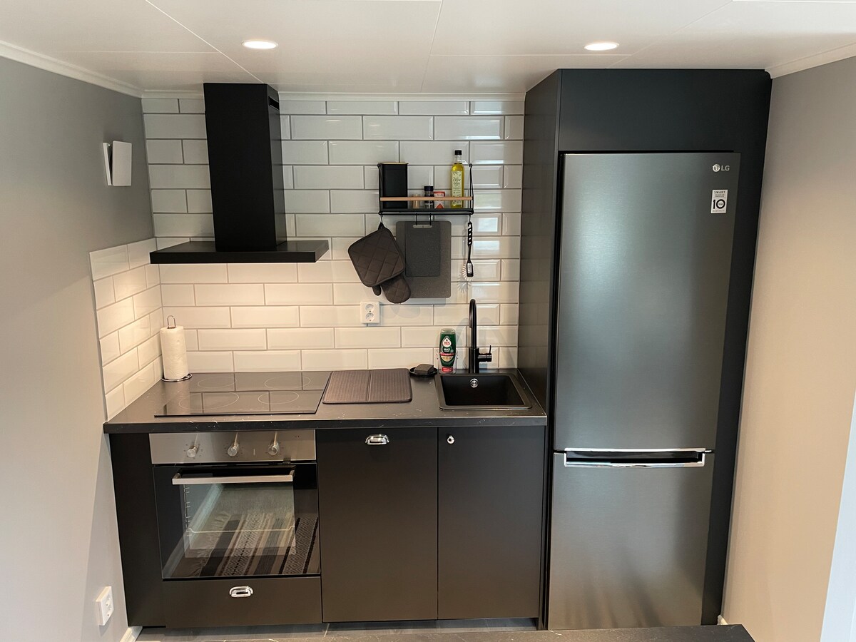 A modern kitchen is presented with sleek black cabinetry and white tiled backsplash. It features a stove, a refrigerator, and a sink, with various cooking essentials neatly arranged on a wall-mounted shelf. Natural light illuminates the space, enhancing its functional design.
