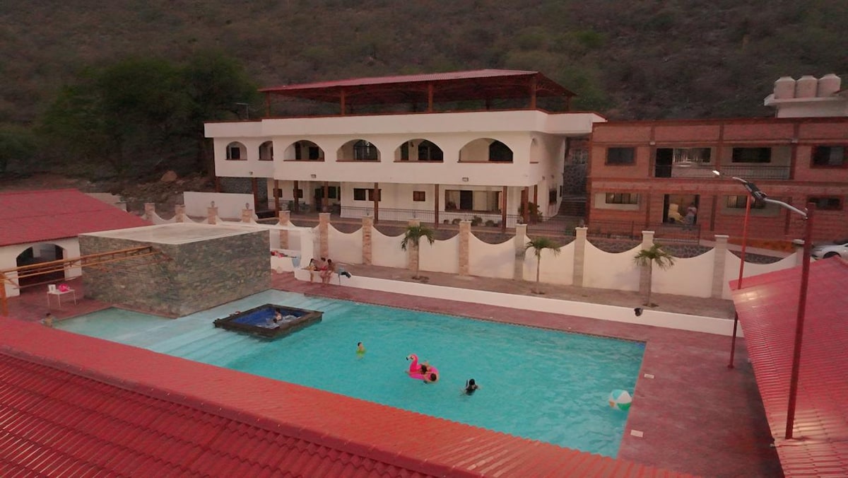 An expansive swimming pool is surrounded by a decorative stone wall and relaxation areas. The two-story building provides shaded outdoor seating. Several guests enjoy the water, with colourful floats present. Lush hills frame the scene, creating a serene setting for leisure activities.