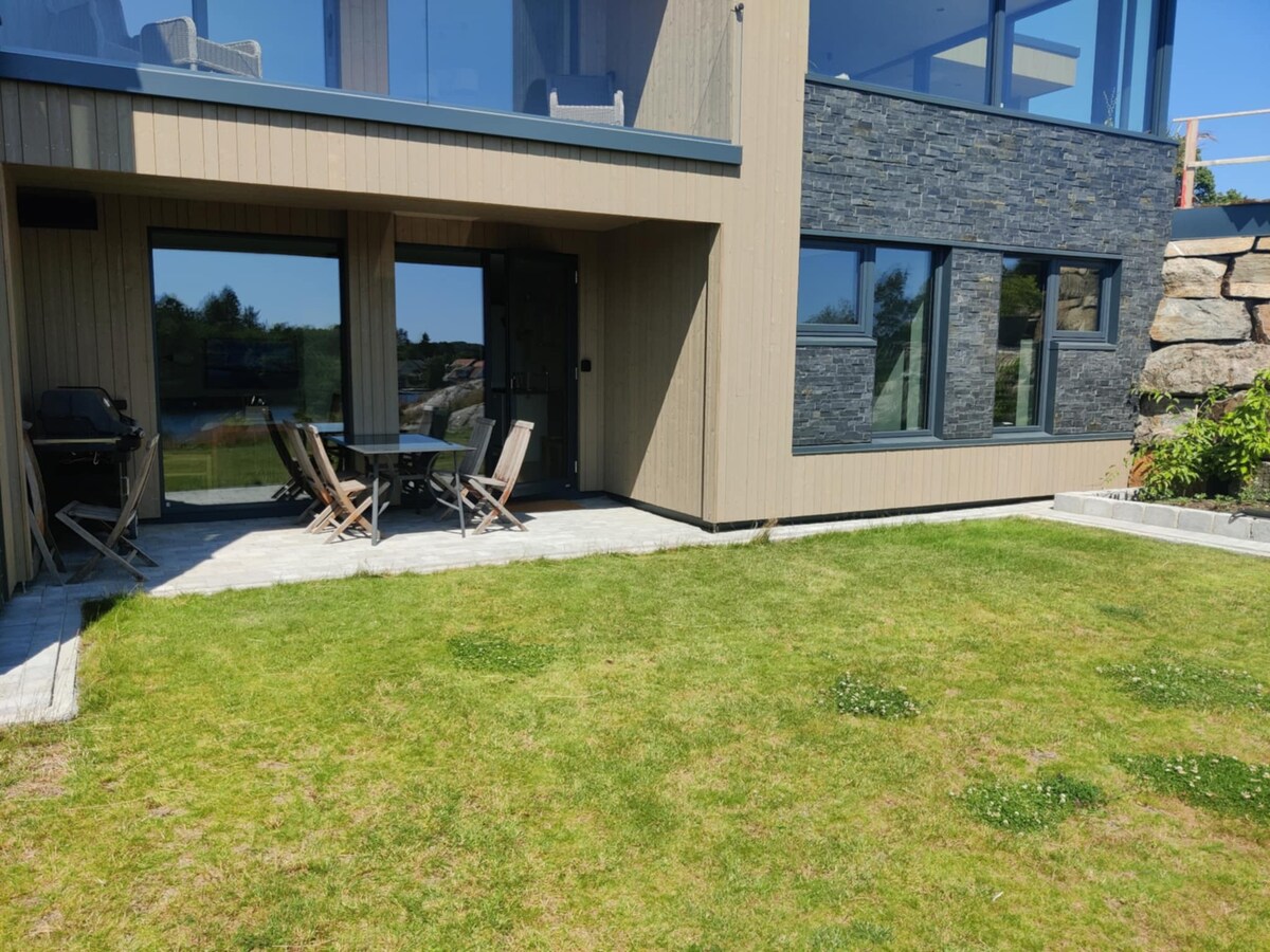 An outdoor patio area is visible, featuring a table surrounded by several chairs. A grill is positioned to one side. The space is complemented by a lush green lawn, offering a tranquil setting with a backdrop of the modern building's exterior.