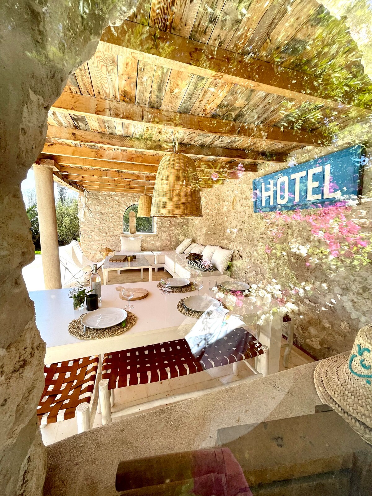 A shaded outdoor seating area showcases a natural wood ceiling supported by stone columns. Comfortable seating is arranged around a table set with plates and decor. A colorful 'HOTEL' sign hangs prominently, with greenery visible through the opening, adding to the tranquil ambiance.