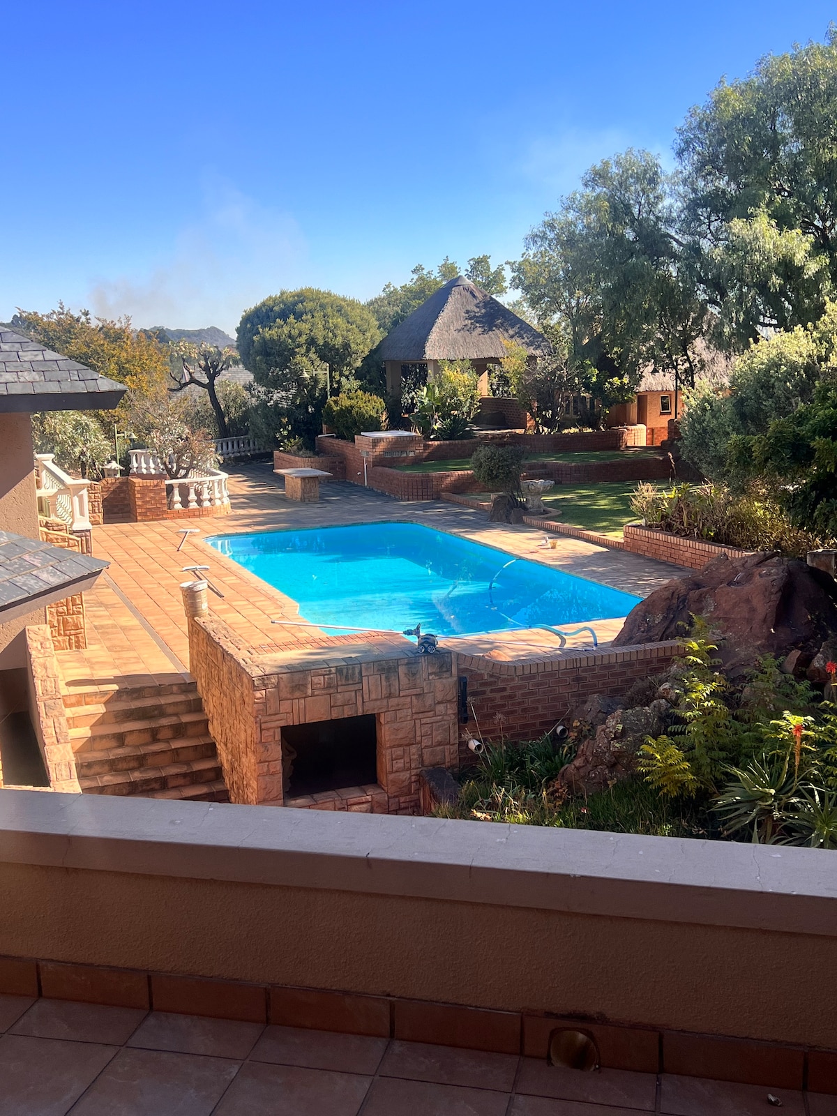 An outdoor area features a large swimming pool surrounded by a well-maintained garden and various trees. A thatched gazebo is visible in the background, along with stone pathways and staircases leading to the poolside. Clear blue skies enhance the serene atmosphere.